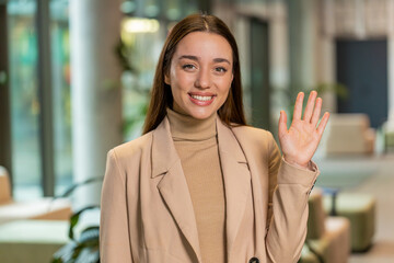 Hello. Happy young Caucasian businesswoman employee waves hand hi gesture greeting welcomes...