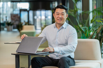 Chinese young businessman closing laptop computer after finishing work at hotel lobby. Smiling...