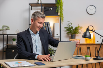 Young gray-haired businessman freelancer in formal suit opens laptop start working sends messages,...