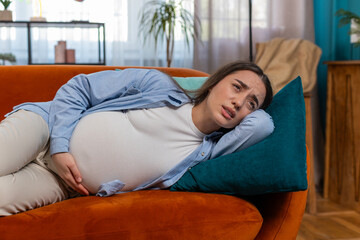 Upset young pregnant woman suffering from abdominal pain lying on couch in living room at home....