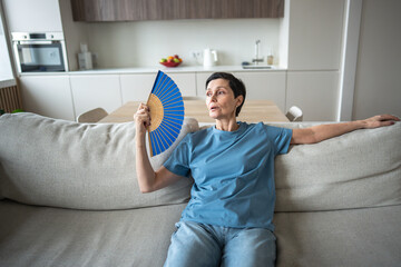 Unwell exhausted mature woman sitting on home couch waving paper fan to cool down. Overheated annoyed female suffers from stuffiness, feeling hot during heatwave in apartment without air conditioning.