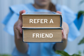 Young girl holding two books with inscription on side: REFER A FRIEND. Referral marketing or affiliate marketing concept. Invite friends to refer a friend loyalty program to win online reward, earn.