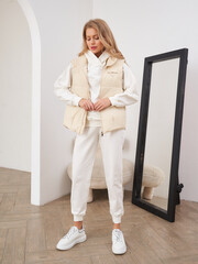 A woman with long hair stands in a stylish outfit, wearing a beige vest over a white hoodie, white...