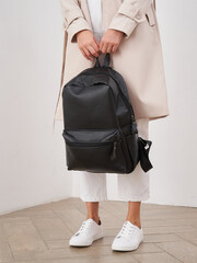 Woman in beige coat and white pants holding a black backpack by the handle, wearing white sneakers,...
