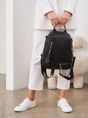 Woman in white outfit holds black backpack with metal accents, standing on wooden floor. White...