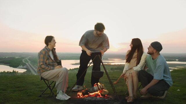 Group of young people are chatting, laughing and smiling during hike. One of them is telling funny jokes and telling stories around campfire as they cook food in pot and relax in nature.