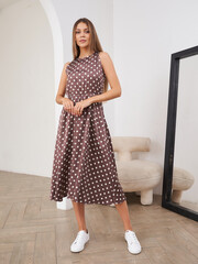 Woman in sleeveless brown polka dot dress stands confidently in room with parquet floor, large...