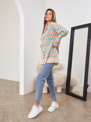 Woman poses confidently in stylish patterned cardigan and jeans, wearing white sneakers. Standing...