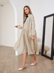 A woman in a flowy beige dress with black polka dots and a black bow detail poses indoors next to a...