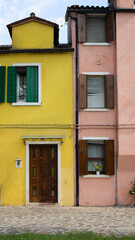 A vivid row of colorful residential houses in Burano, Italy, featuring bright facades in yellow, pink, blue, orange, and peach. Clothes hang to dry between windows, and palm trees grow in. Vertical
