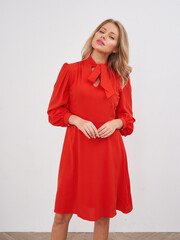 Blonde woman wearing bright red dress with puffed sleeves and tied bow at neck, posing confidently...