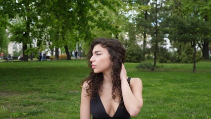 A young woman sits in a vibrant park, basking in the sunlight, her curly hair flowing gently as she enjoys the peaceful atmosphere and greenery around her.
