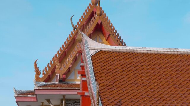 Traditional Thai Buddhist temple with beautiful red tiled roof with magnificent chofas in Thai architectural style on the background of blue sky. Shot in motion 