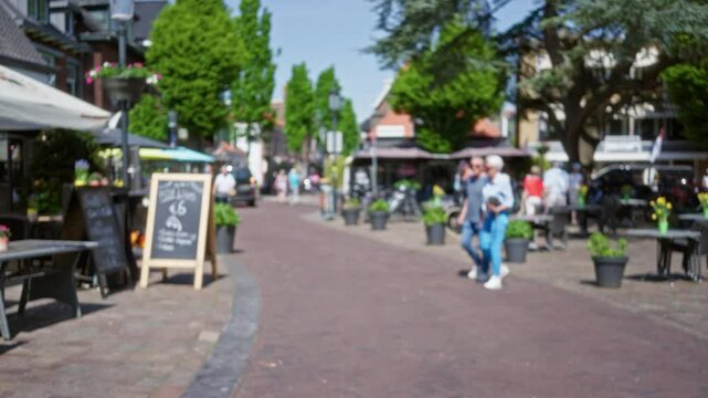 People walking leisurely on charming european city street with outdoor cafes and lush greenery in background on a sunny day reflecting vibrant urban life