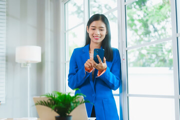 An Asian businesswoman in a blue blazer celebrates with joy while looking at her smartphone. She...