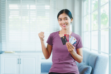 Happy Asian woman sits comfortably on a sofa,smiling while holding multiple credit cards,representing convenient online shopping,smart financial planning,modern digital consumption in a relaxed home