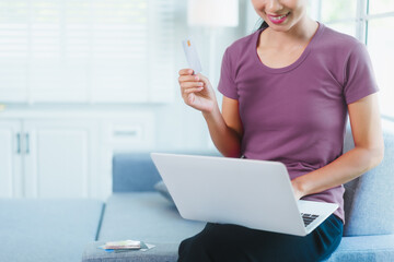 Happy Asian woman sits comfortably on a sofa,smiling while holding multiple credit cards,representing convenient online shopping,smart financial planning,modern digital consumption in a relaxed home
