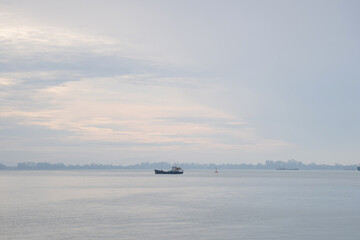 Obraz premium Fishing boat cruising through calm coastal waters under a soft pastel sky during early morning light, with distant shoreline and gentle clouds in the background.