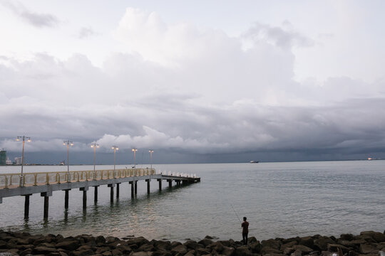 Evening view of a fisherman standing by a rocky shore near a pier with street lamps lit, under dramatic cloudy skies over calm sea waters