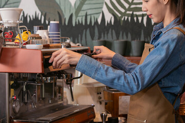 Side view, young attractive Asian barista female using professional coffee machine to make hot...