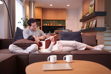 Relaxing Asian Couple Enjoying Leisure Time Together on Couch at Home Reading and Surfing Internet...