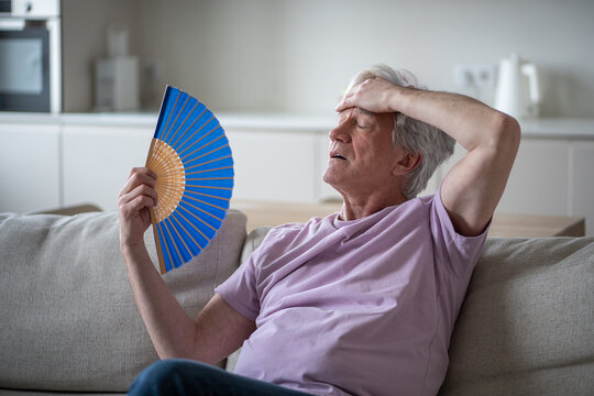 Exhausted annoyed elderly man fanning himself at home, drenched in sweat. No air conditioning, hard to breathe, suffocating, scorching weather, heart strain, heatwave discomfort, intense summer heat