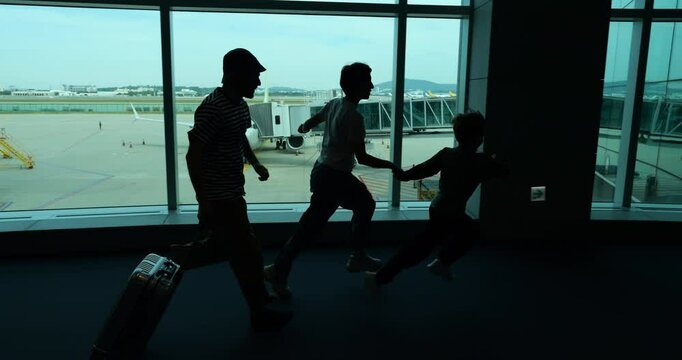 Silhouetted shot of family energetically running through airport terminal, with young boy leading and father with suitcase trailing behind. Passengers hurrying toward boarding gate in funny rush