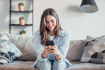 Young woman sitting on sofa at home, using phone with varied emotions happy, sad, angry and...