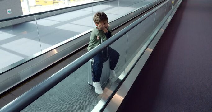 Sad or tired young boy sitting on suitcase, sliding on moving walkway inside airport terminal, displaying exhaustion and boredom after a long flight. Slow motion tracking shot