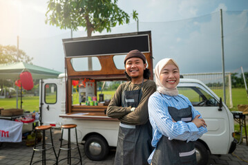 Indonesian southeast asian male and a Chinese muslim female in hijab couple barista standing with confidence in front of their coffee shop. A small business of a coffee shop or cafe on a truck