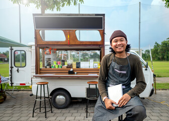 Indonesian southeast asian male barista sitting while holding a tablet in front of his coffee shop. A small business of a coffee shop or cafe on a truck