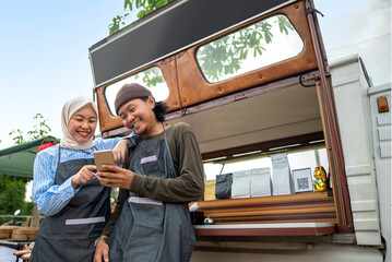Indonesian southeast asian male and a Chinese muslim female in hijab couple of baristas using a cellphone in front of their coffee shop. A small business of a coffee shop or cafe on a truck