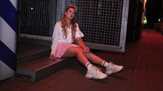 A girl with long blond hair in a pink miniskirt and a white blouse sits in a recreation park at night.