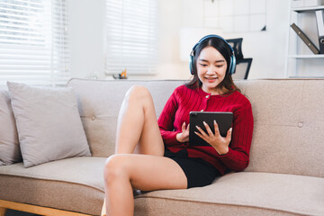Young woman relaxing on sofa with headphones, smiling while using digital tablet in bright living room, casual and comfortable atmosphere, enjoying technology and leisure time