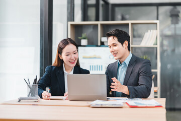 Two Asian business professionals in formal attire collaborate at modern office desk, smiling and discussing work while using laptop, with documents and charts in background