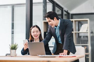 Two Asian businesspeople, woman sitting and man standing, collaborate in modern office, smiling and discussing work at desk with laptop, smartphone, and documents