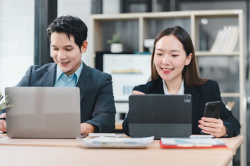 Asian businesspeople working together at office desk, using laptop, tablet, and smartphone, smiling and collaborating on project in modern workplace environment