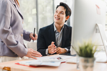 Two business professionals, one man sitting at desk and one woman standing, discuss work in modern office with natural light, documents, and computer, showing positive atmosphere