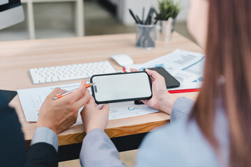Two business professionals discuss data while one holds blank smartphone, sitting at modern office desk with documents.