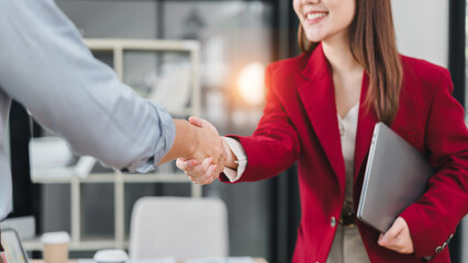 Businesswoman in red blazer smiling and shaking hands with colleague in modern office, holding laptop, teamwork and partnership concept © PRIME STOCK LAB