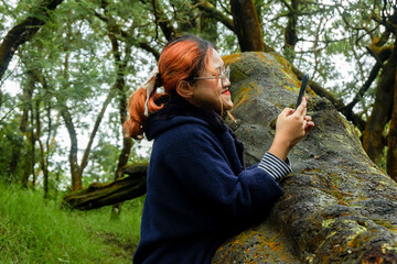 Smiling woman with orange hair and glasses uses her phone while leaning on a mossy fallen tree in a peaceful forest. Concept of relaxation, digital lifestyle, and nature.........
