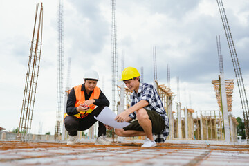An architect and engineer discuss blueprints on a construction site, wearing helmets, ensuring safety, quality, and progress through collaborative planning.