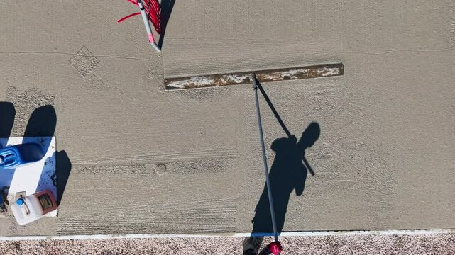 Drone video of worker leveling concrete slab with long trowel Aerial video showing the shadow of a construction worker using a long-handled trowel to smooth freshly.