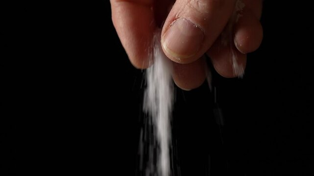 Male hand sprinkling or adding salt on black background to dish in kitchen, cooking food, slow motion. Chef adds spices