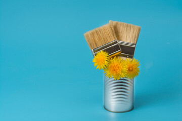 Repair brushes and yellow dandelions gracefully arranged in a paint can on a blue background. Spring and summer repair concept. Copy space