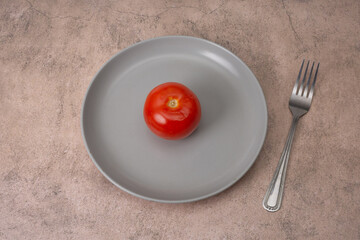 Red tomato lies on a gray plate, a fork is nearby. Strict diet for weight loss. Flat lay
