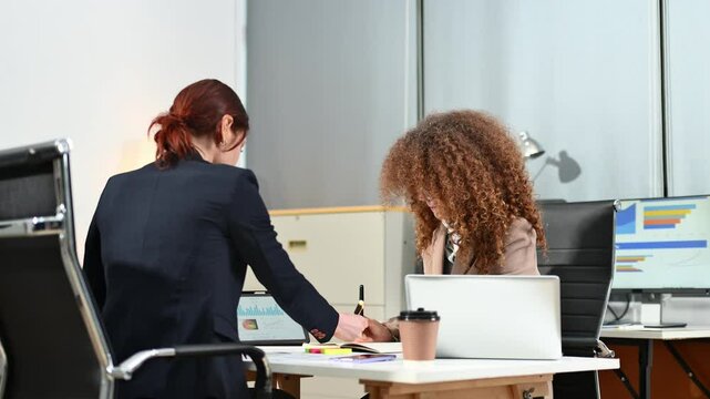 Two businesswomen smiling and discussing data charts in a bright modern office. Collaboration, startup culture, and teamwork