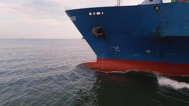 Side close-up view of a large blue cargo ship moving through the sea, showing its bow, anchor, and visible red hull below the waterline. Maritime transport and international shipping concept