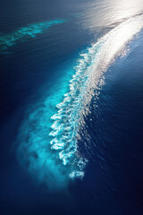 Aerial view of a boat's wake traversing deep blue ocean waters, creating a vibrant contrast of white foam against the dark blue, with hints of lighter turquoise shallows visible beneath the surface
