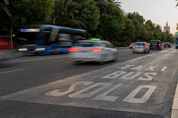 A busy city street at dusk with blurred moving vehicles showing taxi and bus lane markings on the...
